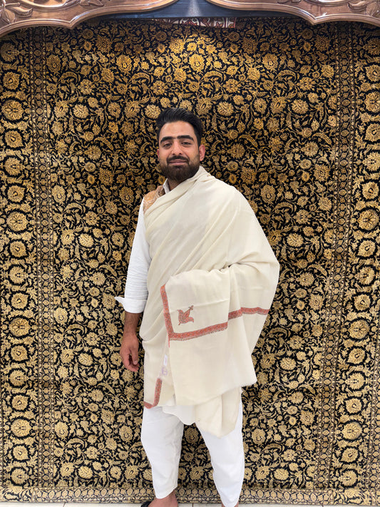 Man in traditional white attire standing against a patterned wall.
