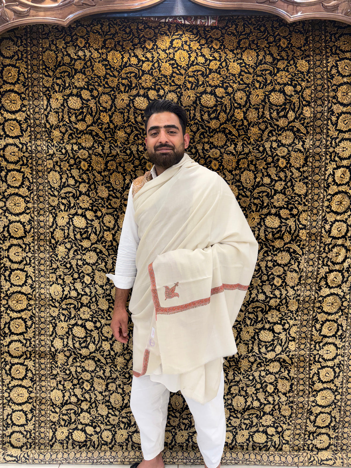 Man in traditional white attire standing against a patterned wall.