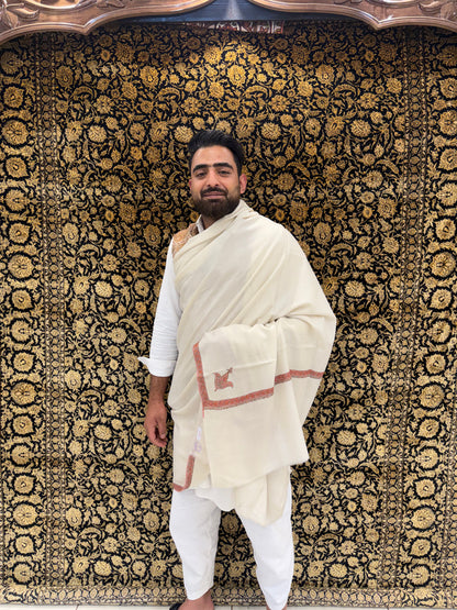 Man in traditional white attire standing against a patterned wall.