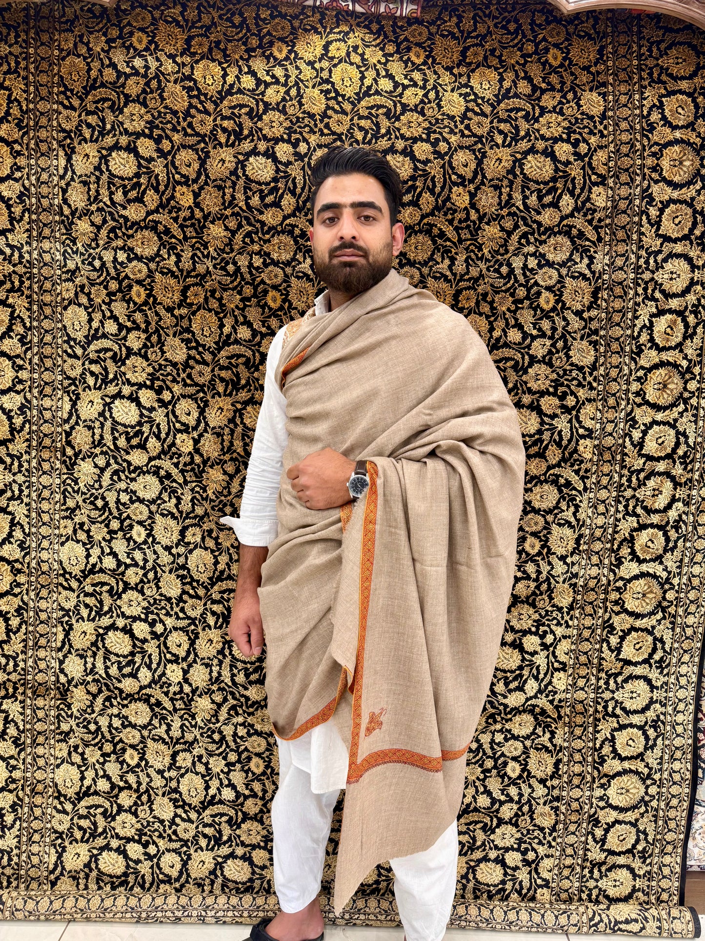Man wearing a beige shawl with orange border against a patterned wall.