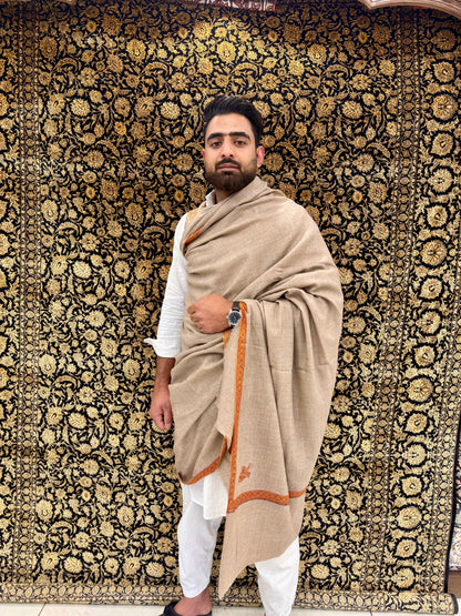 Man wearing a beige shawl with orange border against a patterned wall.