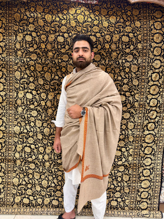 Man wearing a beige shawl with orange border against a patterned wall.