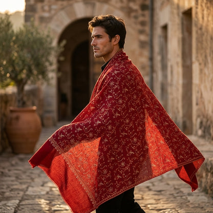 Man wearing a red patterned shawl in an outdoor setting with stone architecture and potted plants.